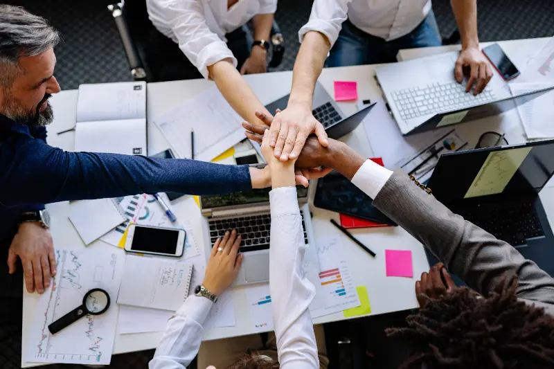 A team joining hands over a desk covered in plans and laptops